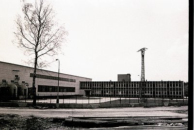 Mid-century industrial building with Brutalist concrete façade and large grid windows, flanked by a lone tree and railway tra...
