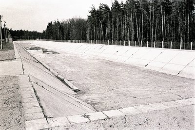 Concrete pathway flanked by parallel trenches, bordered by a fence and leafless trees. Likely a former concentration camp or ...