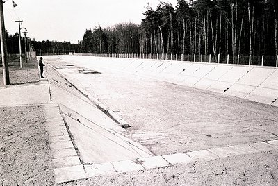 Concrete platform with deep grooves, flanked by barbed-wire fencing and young trees. A lone figure stands near the entrance, ...