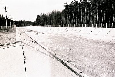Concrete race track with grooved surface, flanked by gravel and bordered by a fence. Young forest of evenly spaced trees in b...