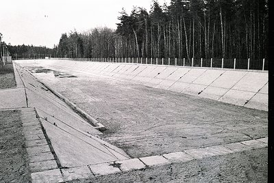 Empty concrete pathway flanked by barbed-wire-topped fences, flanked by dense forest. Likely a WWII-era concentration camp or...