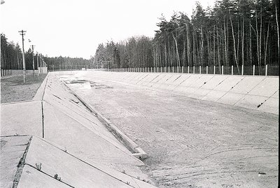 Empty concrete stadium with tiered seating, likely mid-20th century. Rows of wooden poles line the perimeter, possibly for li...