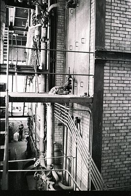 Industrial brick factory interior with mid-century mechanical equipment. Twin vertical pipes and metal scaffolding dominate t...