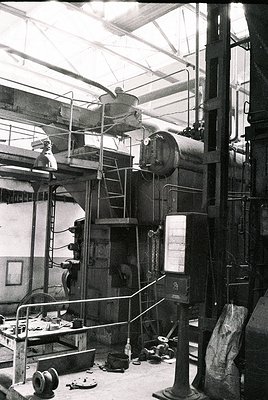 Industrial steam-powered machinery in a mid-20th century factory setting. Visible components include a large boiler, steam pi...