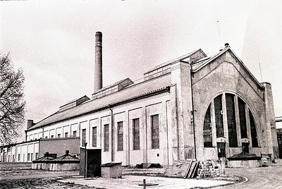 Industrial-era brick building with Art Deco architectural elements, featuring a tall chimney and arched entrance. Signage sug...