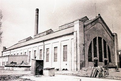 Industrial-era brick building with Art Deco-inspired arched entrance and tall chimney stack. Exterior shows signs of aging an...