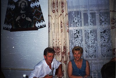 Vintage interior featuring two individuals seated at a table, framed by intricate lace curtains and a religious icon depictin...