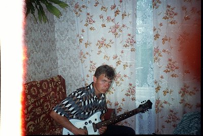 Vintage indoor portrait of a man playing a white electric guitar in a dimly lit room with floral wallpaper. His striped shirt...