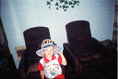 Vintage indoor portrait of a young child wearing a wide-brimmed hat with floral patterns and a colorful, cartoon-themed shirt...