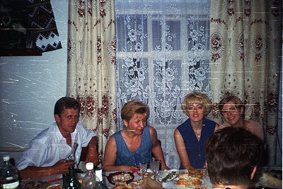 Vintage indoor gathering featuring four adults seated at a table with patterned lace curtains and floral wallpaper. Mid-centu...
