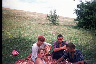 Vintage outdoor portrait of three individuals seated on grassy hillside, likely 1960s-1970s. One person’s hair is being style...