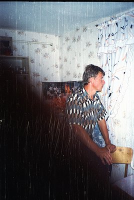Man stands in a shower with diagonal-striped shirt and blue pants, leaning on a wooden ledge. Floral-patterned shower curtain...