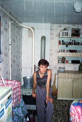 A woman in a patterned sleeveless top and wide-legged pants stands in a modest kitchen, leaning against a stove with a metal ...