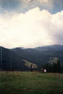 Three figures stand on a grassy hilltop under dramatic, rain-soaked skies, framed by vertical rain streaks. Lush forested val...