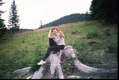 Vintage candid of person posing on a stump in a forested hillside, mid-1990s. Casual attire includes a light jacket, dark pan...