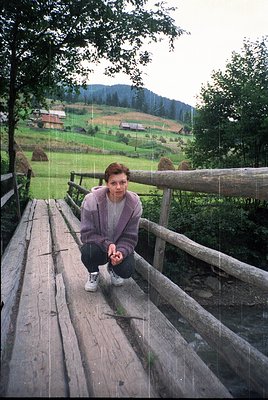 A person sits on a rustic wooden bridge over a stream, surrounded by lush green meadows and rolling hills. The individual wea...