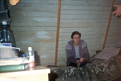 Indoor shot of a person seated on a camouflage-patterned bed in a rustic wooden cabin. Bottles of water and a plastic contain...