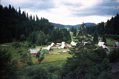 Aerial view of a rural campsite nestled in a forested valley, likely mid-20th century. Several pitched tents and a few small ...