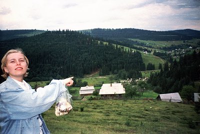 A woman in a denim jacket and light blue shirt poses on a grassy hillside, pointing toward a valley with dense forest and sca...