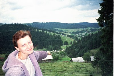 A woman poses in a lush, forested valley, pointing toward a distant village. She wears a purple sweater over a white top, set...