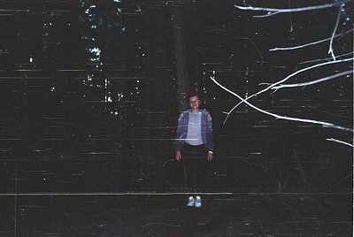 Portrait of a person standing under illuminated tree branches against a dark, textured wall. Light trails create dynamic patt...