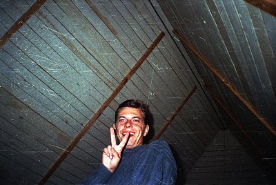 Vintage indoor shot of a man in a blue sweater making a peace sign under a rustic wooden ceiling with exposed beams. Likely 1...