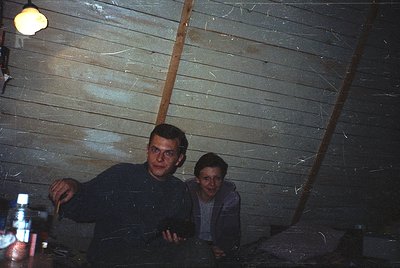Vintage indoor shot of two men in a rustic wooden cabin, likely 1970s–1980s. Dark, textured planks and a single bulb illumina...