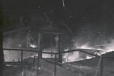Vintage black-and-white photo of a **mining or industrial site** at night, featuring a **suspended platform** with two worker...