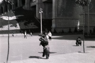 Vintage black-and-white street scene featuring a woman in a long, flowing dress walking with a child in a carrier. Urban sett...
