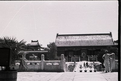 Black-and-white photograph of traditional East Asian temple courtyard, featuring tiled roofs, stone pathways, and potted plan...