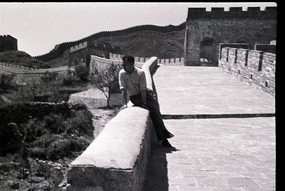 Black-and-white shot of a lone figure perched on a stone barrier atop the Great Wall of China, mid-gesture. Stone watchtowers...