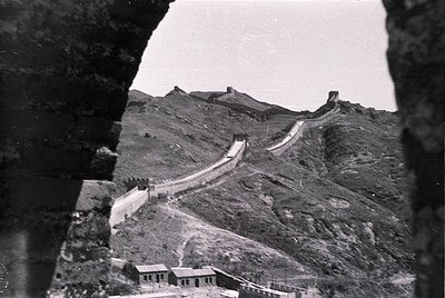 Black-and-white shot of the Great Wall of China winding through rugged terrain, framed by a stone arch. Mid-20th century cons...