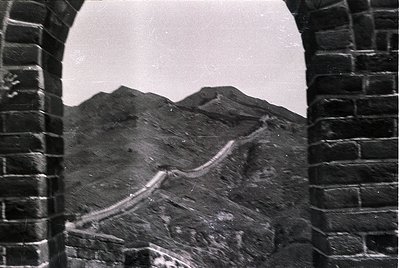 Black-and-white archival shot of the Great Wall winding through rugged mountains, framed by a stone archway. Distinctive bric...