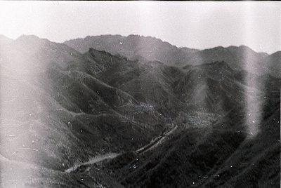 Vintage black-and-white aerial view of rugged mountain terrain with winding road cutting through steep slopes. Dense forest c...