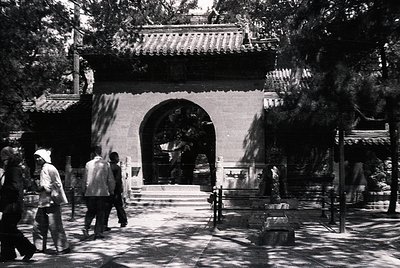 Traditional Chinese temple entrance with curved, tiled roof and stone archway. Visitors in 1970s-era clothing walk through sh...