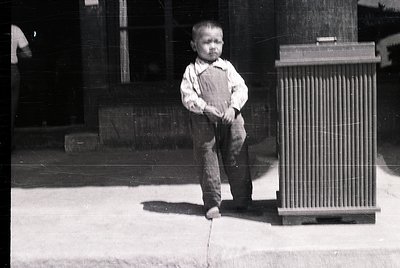 Young boy in early 20th-century attire—overalls, short-sleeved shirt, and sandals—standing on a concrete surface beside a ver...