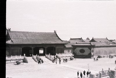 Black-and-white photograph of traditional East Asian palace courtyard with tiled roofs and ornate gateways. Uniformed guards ...