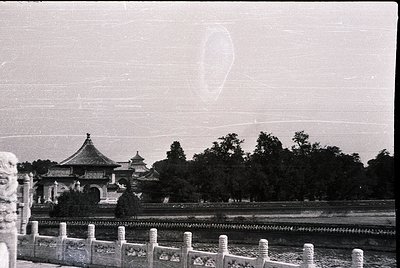 Vintage black-and-white photo of traditional Chinese pavilion with upturned eaves, set beside a stone bridge and lush greener...