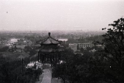 Classic Chinese pavilion with upturned eaves and curved roof ridges, framed by a winding path. Urban sprawl and water body in...