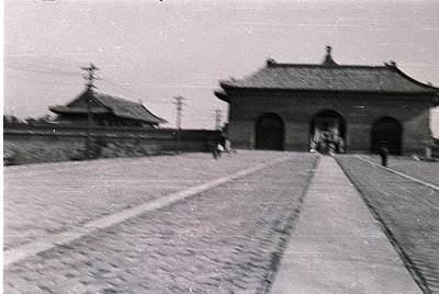 Vintage black-and-white photo of traditional East Asian gate architecture with curved, tiled roofs and arched entryways. Pede...