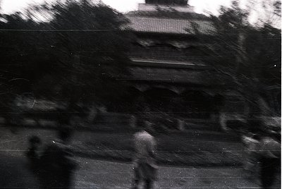 Vintage black-and-white shot of a blurred urban street scene, likely mid-20th century. Silhouetted figures walk along a paved...