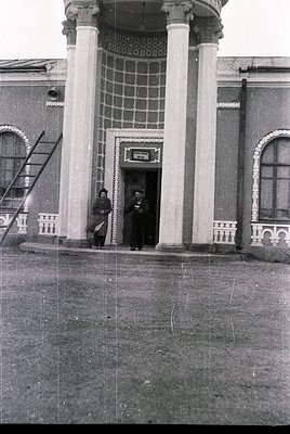 Neoclassical building entrance with two men in early 20th-century attire, standing under a portico supported by fluted column...