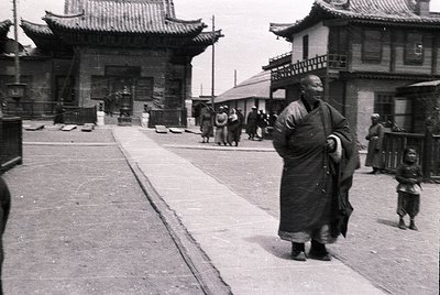 Monk in traditional robe strides along paved street flanked by traditional East Asian architecture with tiled roofs and woode...