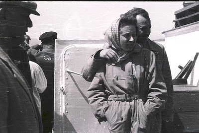 Black-and-white photo of three individuals on a ship’s deck, mid-20th century. A woman in a heavy coat and headscarf leans on...