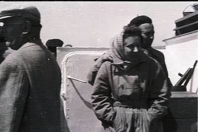 Vintage black-and-white photo of two individuals in heavy winter attire near a military vehicle, likely mid-20th century. The...