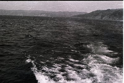 Black-and-white maritime scene showing turbulent waters with visible wake from a vessel. Distant shoreline with forested hill...