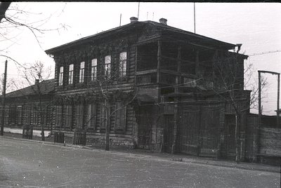 Two-story wooden building with weathered log construction, featuring large rectangular windows and decorative wooden trim. Ex...