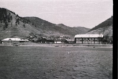 Mid-20th century alpine village nestled between forested slopes. Wooden chalets with gabled roofs line a flat valley floor, s...