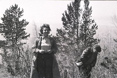 Black-and-white portrait of two individuals in a forested, hilly landscape, mid-20th century. Woman in long coat and headscar...