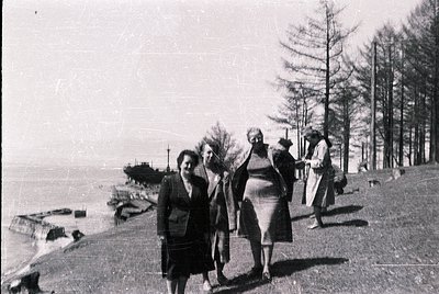 Black-and-white coastal scene featuring four women in 1930s–1940s attire walking along a seaside path. Prominent details incl...
